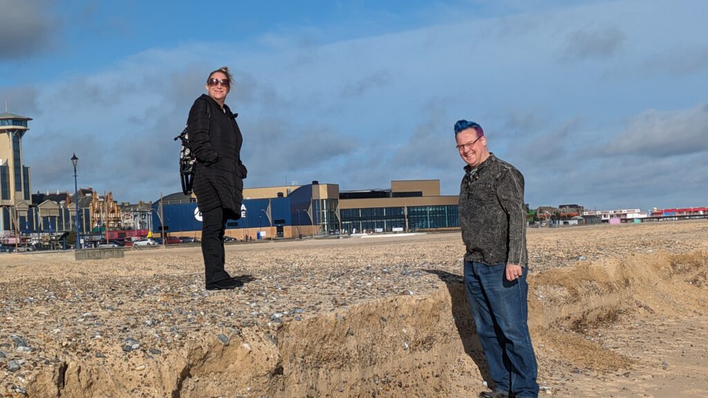 Two people standing on a beach. A woman on the left is on a higher level of sand than the man on the right.